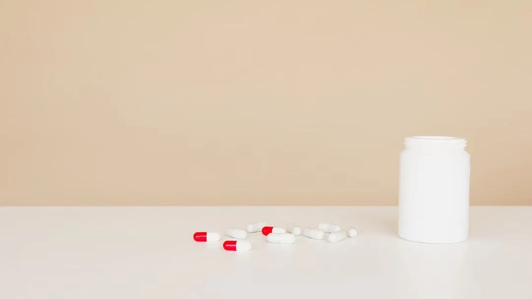 Unbranded white supplement bottle beside loose red and white capsules.