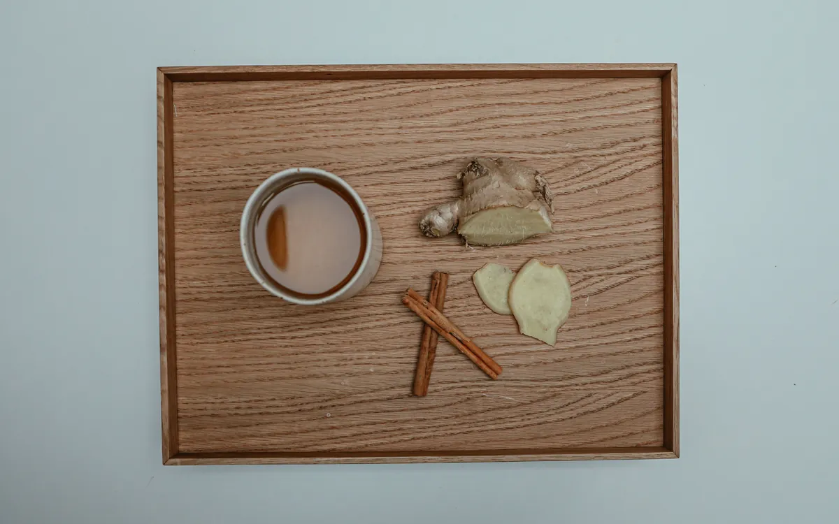 A cup of tea on an oak tray with a piece of fresh ginger root, two ginger slices and two cinnamon sticks.