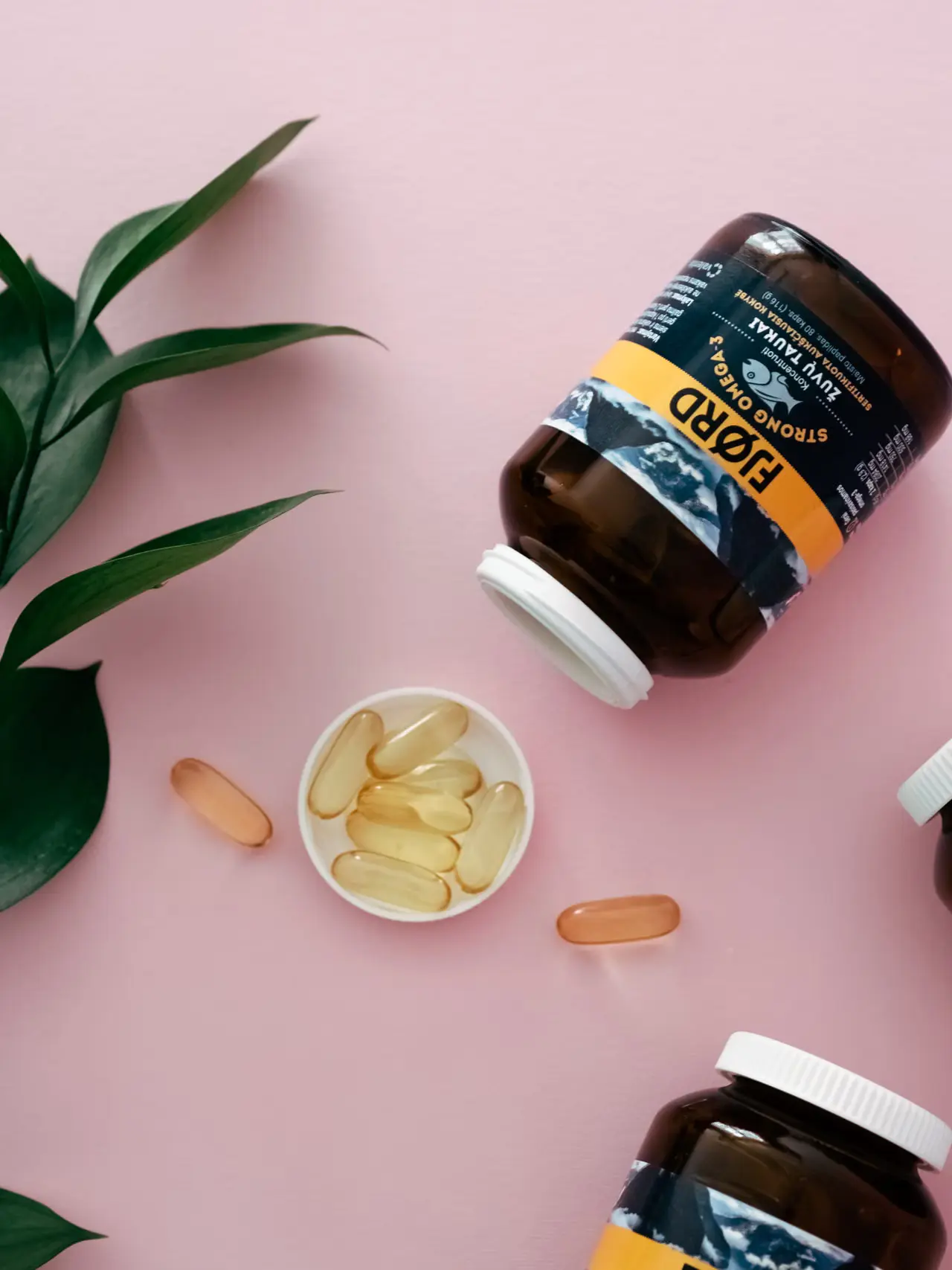 Supplement bottles and capsules with green leaves on a pink surface